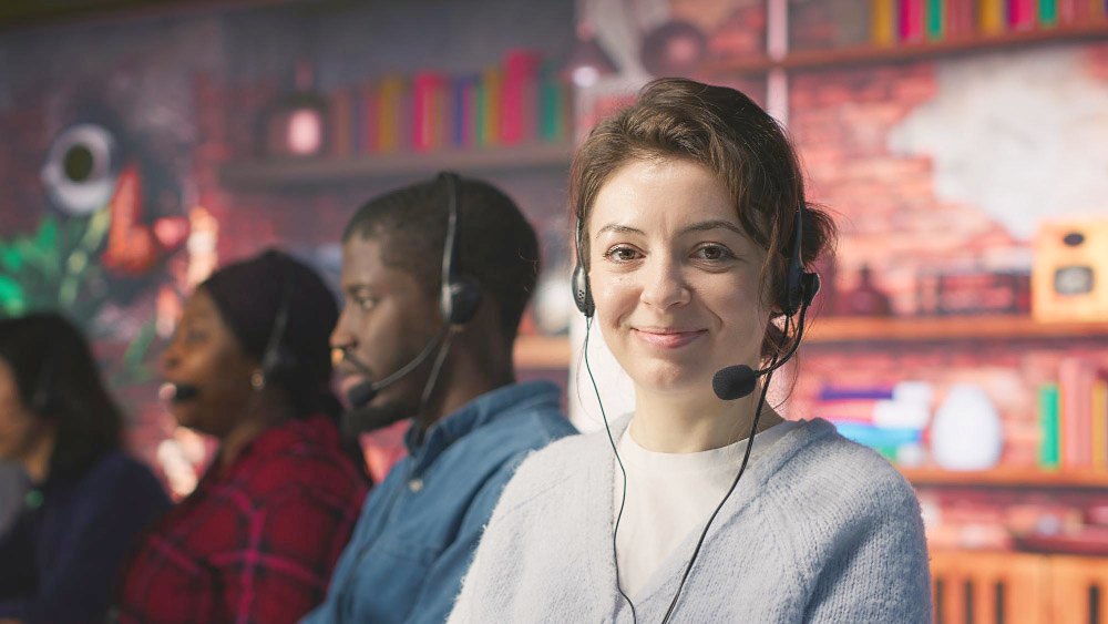 A smiling support agent wearing a headset, with colleagues on calls behind her